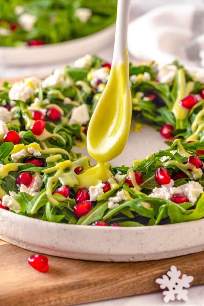 avocado dressing on a spoon being drizzled over a salad on a white plate with pomegranate seeds on top.