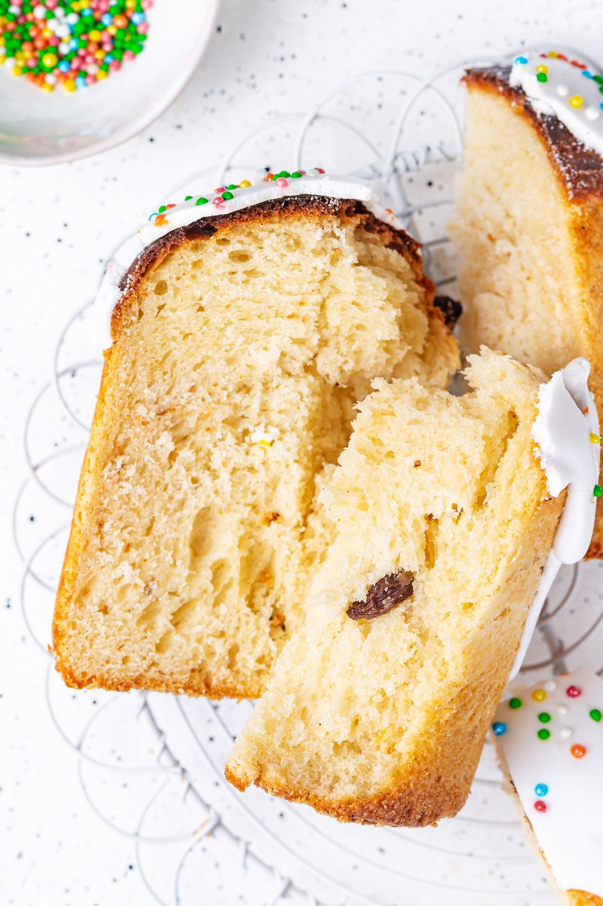 Easter bread cut with frosting and rainbow sprinkles on white surface.