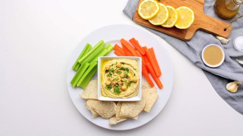classic-hummus-in-a-white-bowl-on-a-white-plate-with-chips-carrots-and-celery-on-it-along-with-a-grey-towel-with-ingredients-a-wooden-board-and-lemon-on-it