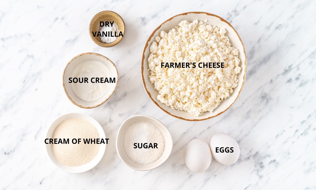 farmers cheese in a bowl and two white large eggs fine white sugar in a white bowl and cream of wheat in a shallow white bowl and sour cream  in a small bowl.