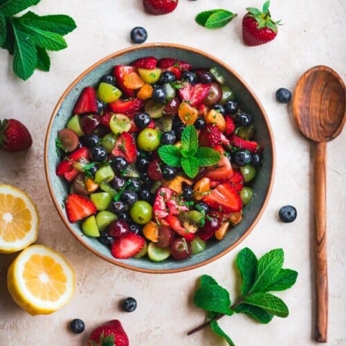 Fruit salad in a bowl with a wooden spoon and fresh fruit around.