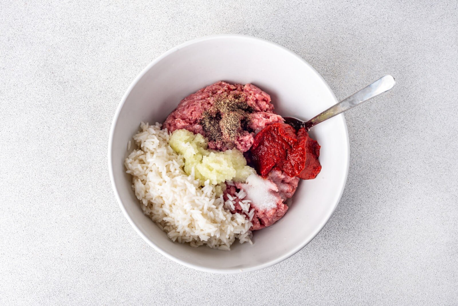 Ground chicken with salt, rice, onion, and pepper in a white bowl with a spoon.