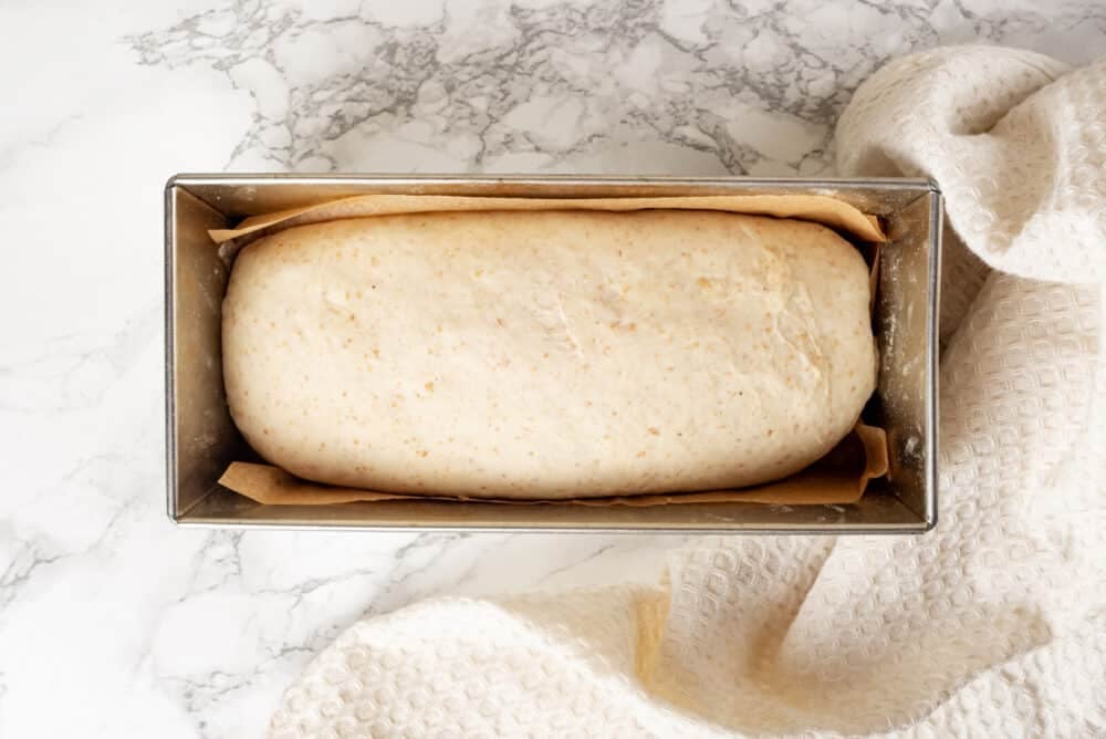 Loaf of bread in a loaf pan with parchment paper, and a towel on the side.