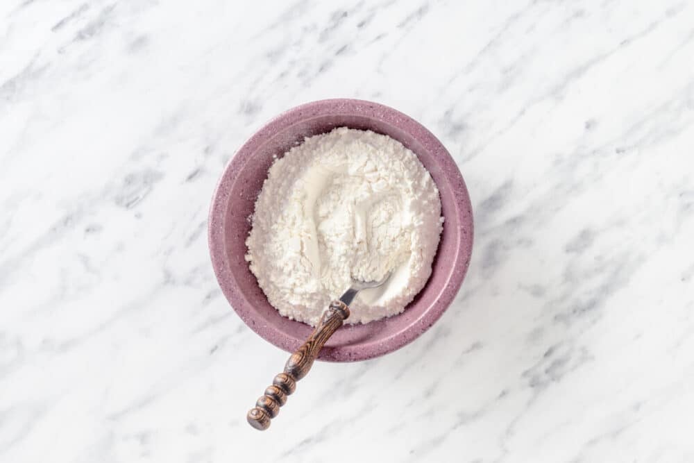 Two flours combining in a purple bowl with a spoon.