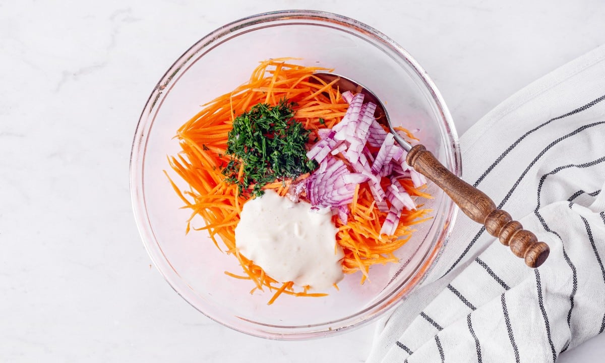 clear medium bowl with grated carrots fresh dill red onion and mayonnaise with wooden spoon.
