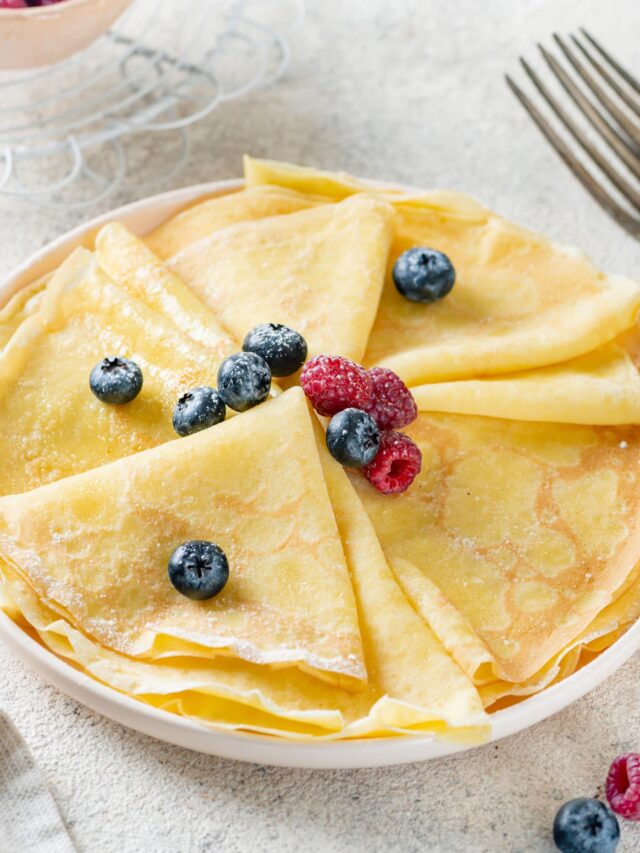 A white plate with folded crepes on it sprinkled with powdered sugar and berries.