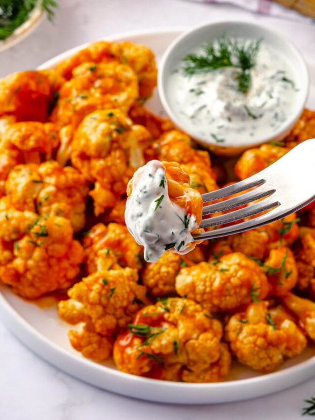 cauliflower florets backed with buffalo sauce on a white plate with yogurt dipping sauce and one floret on a silver fork with some sauce.