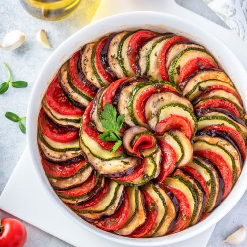 Ratatouille in a round casserole dish on a cutting board surrounded by ingredients for the dish.