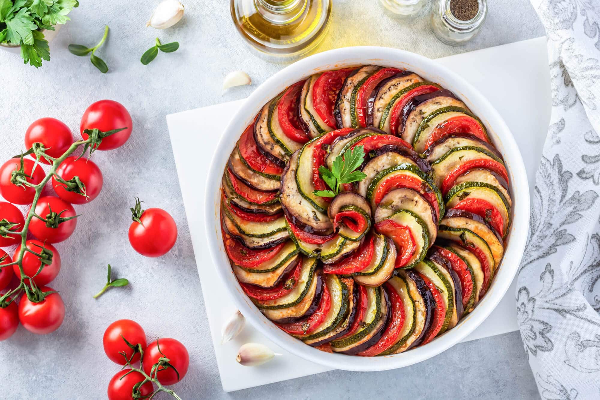 Ratatouille in a round casserole dish on a cutting board surrounded by ingredients for the dish. 