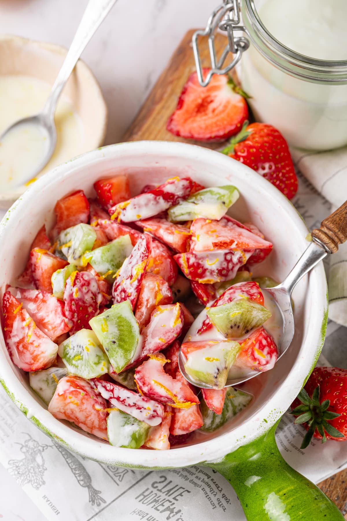 chopped strawberry and kiwi mixed with yogurt orange zest in a white bowl with green handle and wooden stainless steel spoon. 