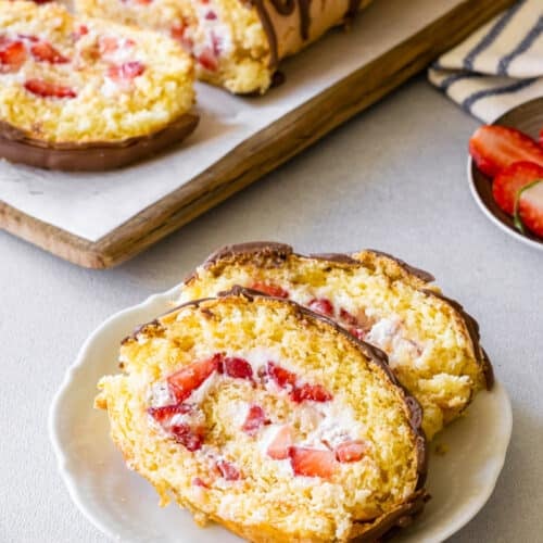 Strawberry cake roll slices on a plate with more cake roll in the background.