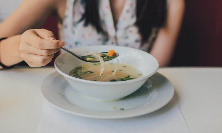 person-eating-soup-at-a-table-from-a-white-bowl-on-a-white-plate-with-a-spoon