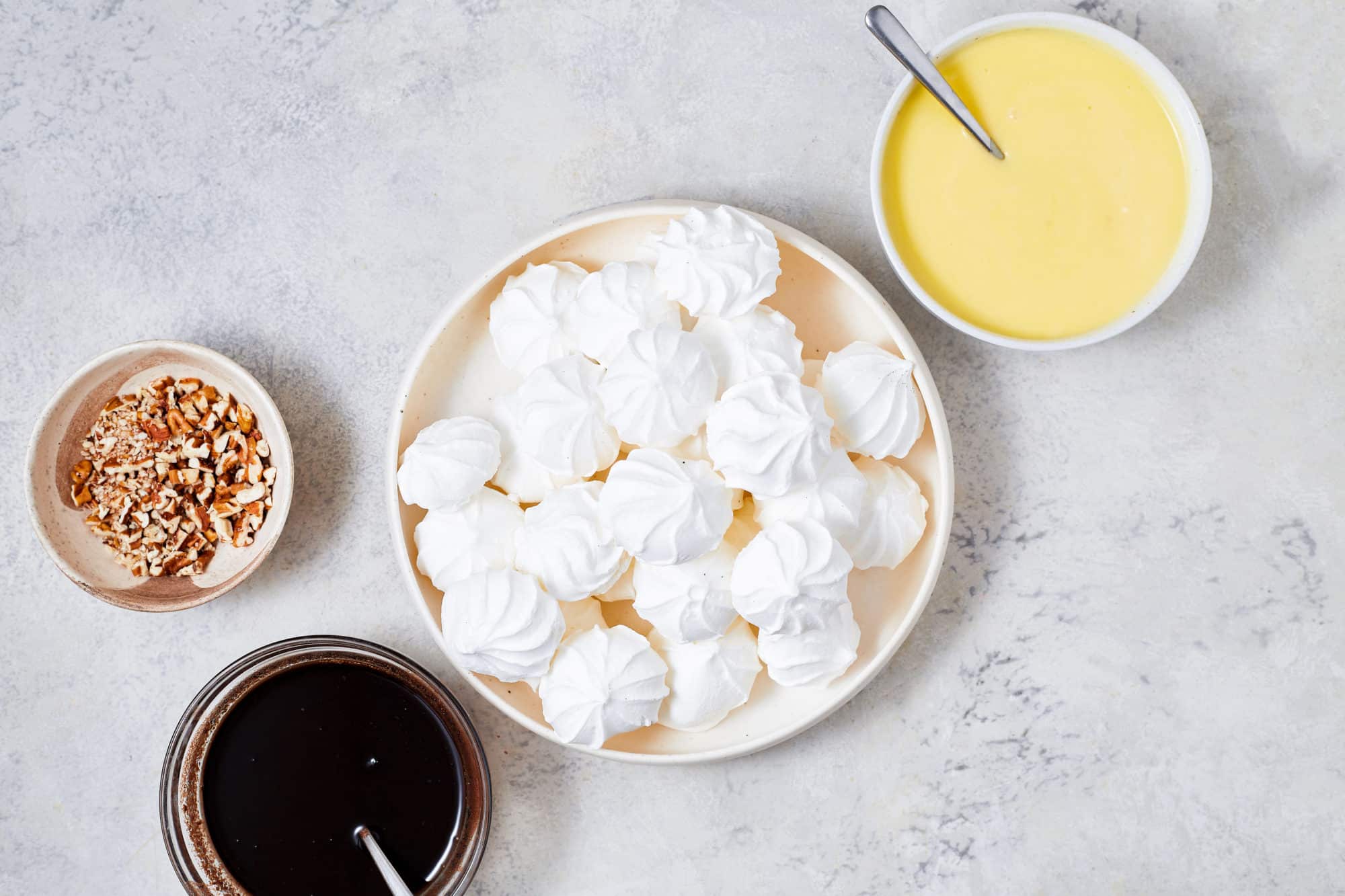 a tan plate filled with meringues with separate dishes on the side filled with custard cream another with melted chocolate glaze and a third with chopped pecans.