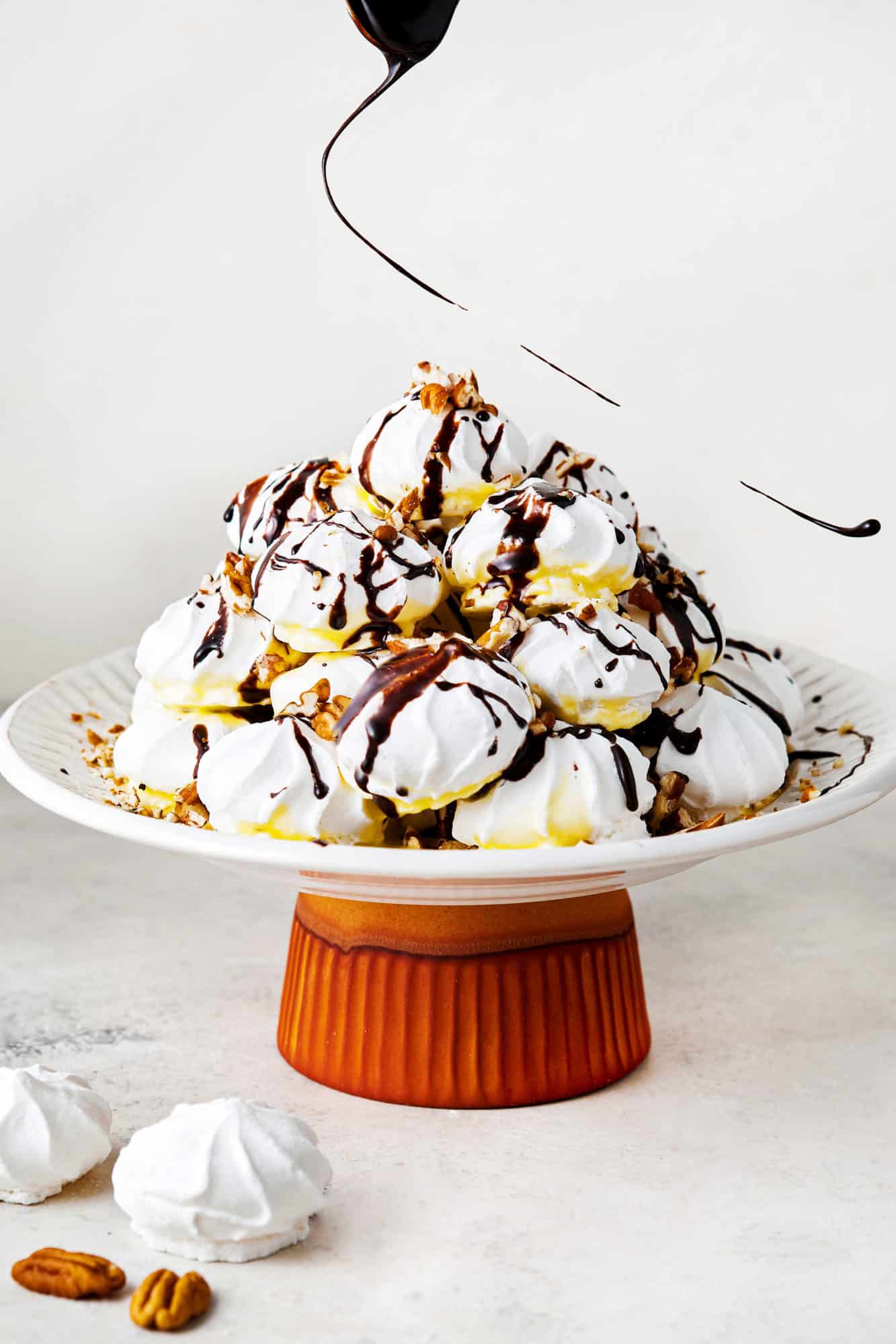 chocolate being drizzled onto a meringue stack shaped like a pyramid on a white plate atop a red ramekin.