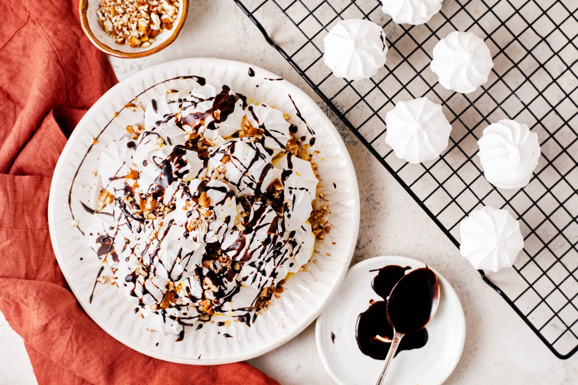 a stack of meringues on a white plate with each layer drizzled in custard cream chocolate glaze and chopped pecans with additional meringues chopped pecans and chocolate on the side on a wire rack and an orange towel.