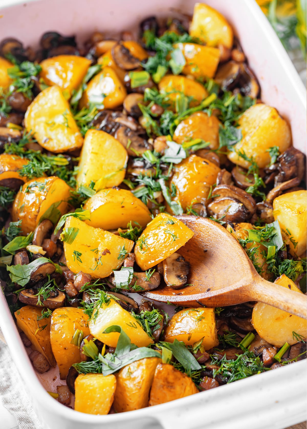 roasted yukon potatoes with mushrooms and pancetta and fresh garnished herbs in a white oven dish and a wooden spoon.