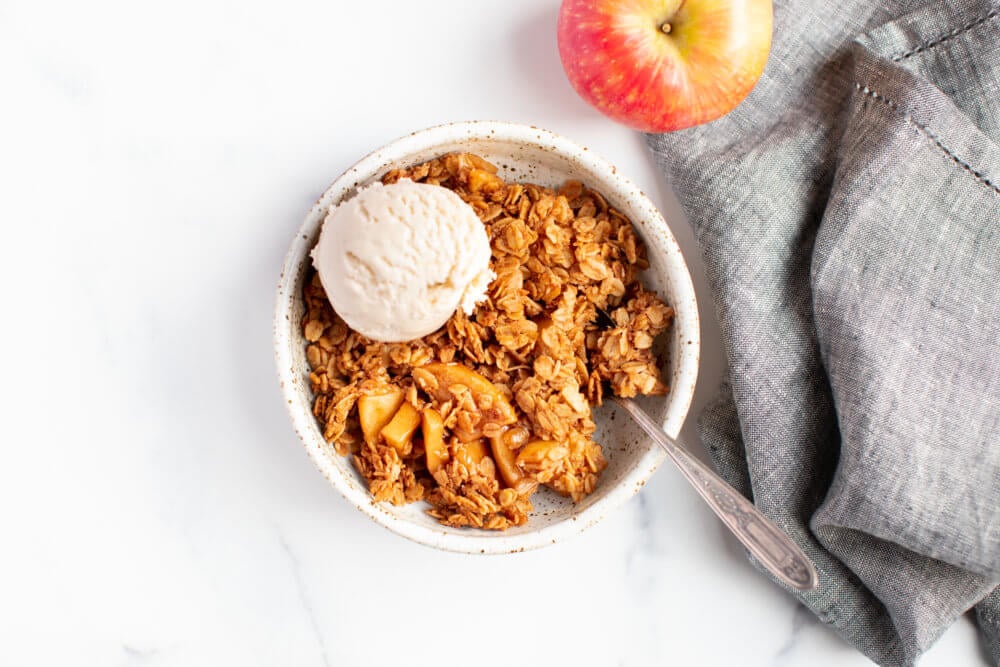 apple crisp in a white bowl with a spoon and a scoop of icecream on top with an apple and a grey towel on the side.