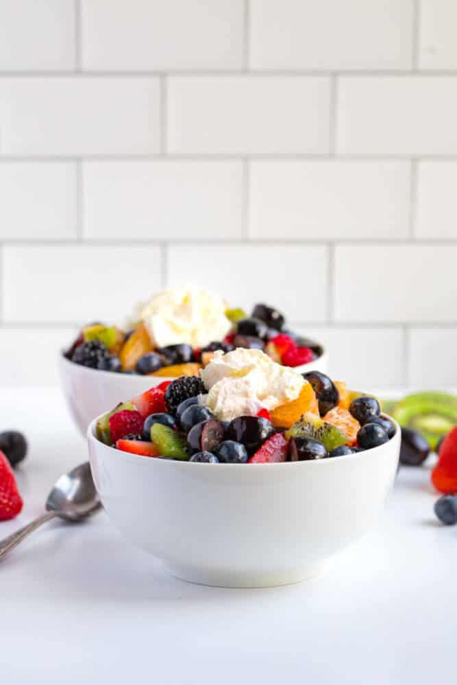 fruit-salad-in-a-white-bowl-with-whipping-cream-on-top-and-a-spoon-on-the-side-and-fruits-in-the-background