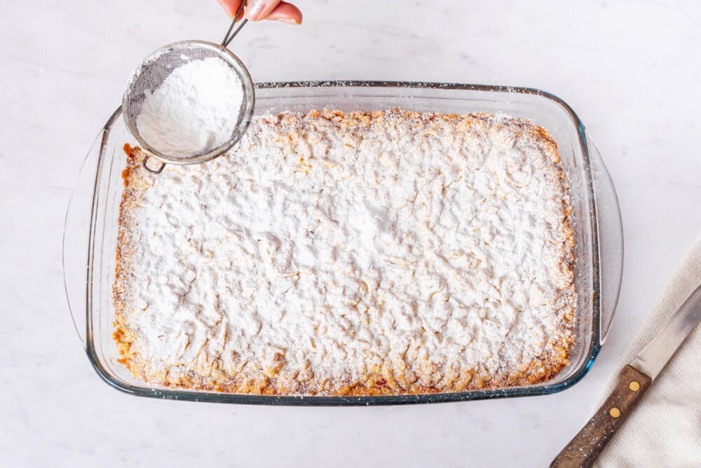 crumbled-jam-bars-in-a-glass-baking-dish-with-powdered-sugar-being-sprinkled-on-top-with-a-knife-on-the-side
