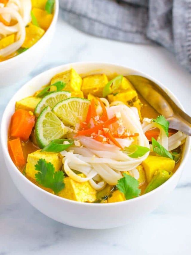 curry noodle soup in a white bowl with a spoon and lime wedges on top and a grey towel on the side.