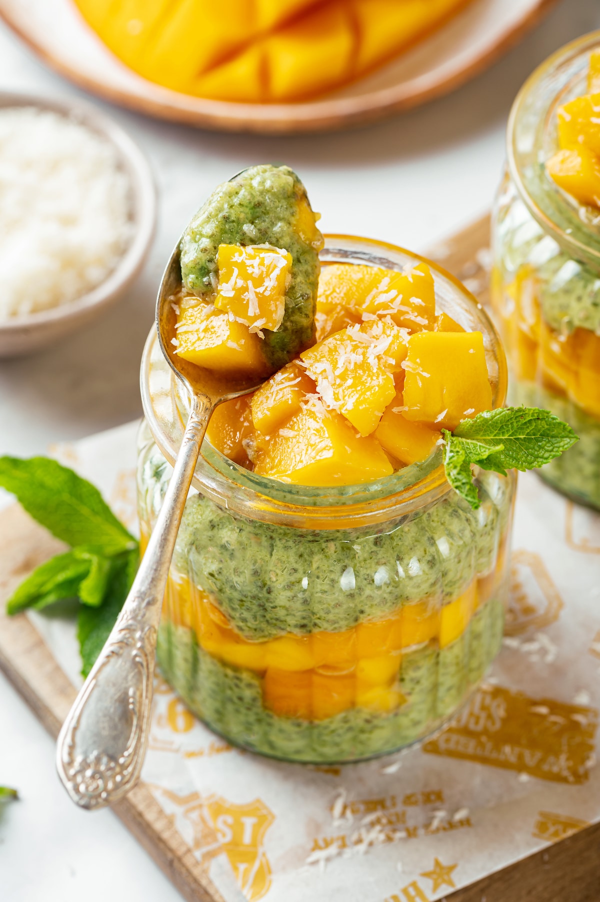 mango and spinach chia seed pudding in two jars with shredded coconut on the top, on a wooden board and parchment paper and silver spoon.