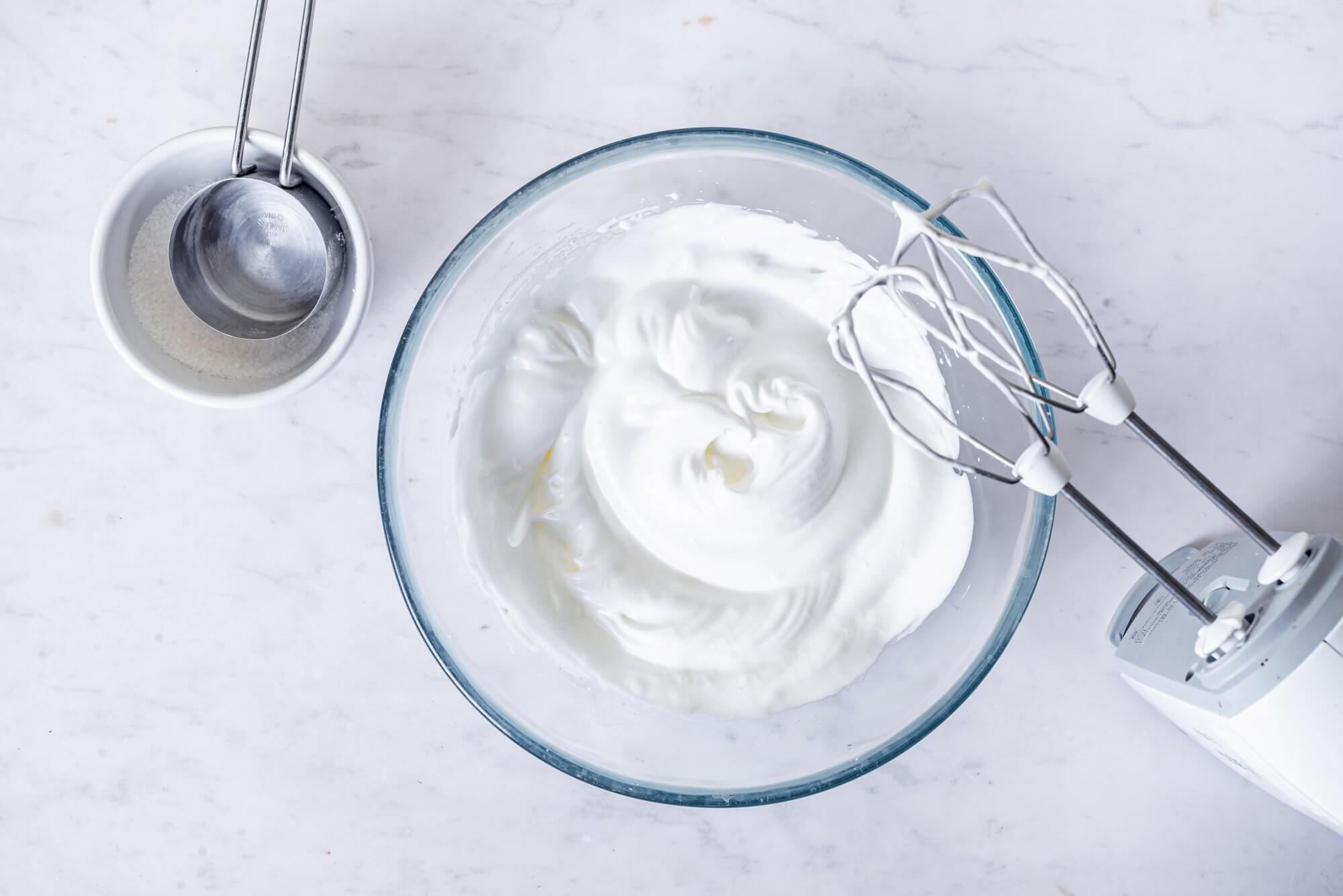 whipped-egg-whites-in-a-glass-bowl-with-hand-mixers-and-a-white-bowl-with-a-silver-measuring-cup-on-the-side