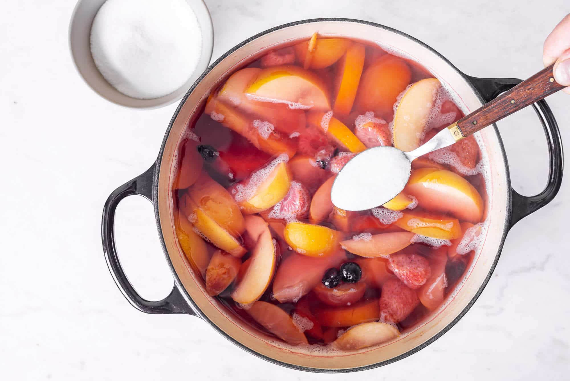 fruits-and-water-in-a-pot-with-sugar-being-added-on-a-spoon-with-a-white-bowl-of-sugar-on-the-side