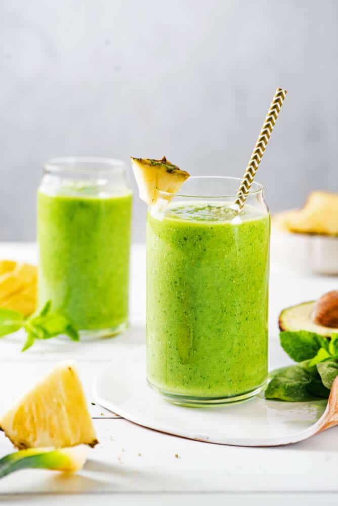 citrus-green-smoothie-in-a-glass-with-a-straw-and-pineapple-wedge-with-ingredients-in-the-background