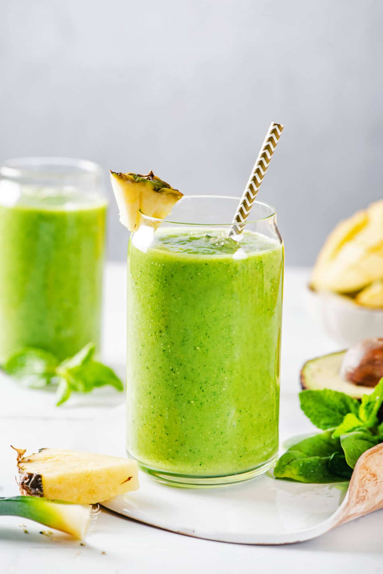 citrus-green-smoothie-in-a-glass-with-a-straw-and-pineapple-wedge-with-ingredients-in-the-background