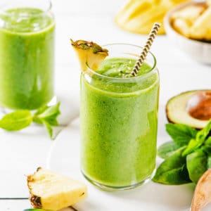 citrus-green-smoothie-in-a-glass-with-a-straw-and-pineapple-wedge-with-ingredients-in-the-background