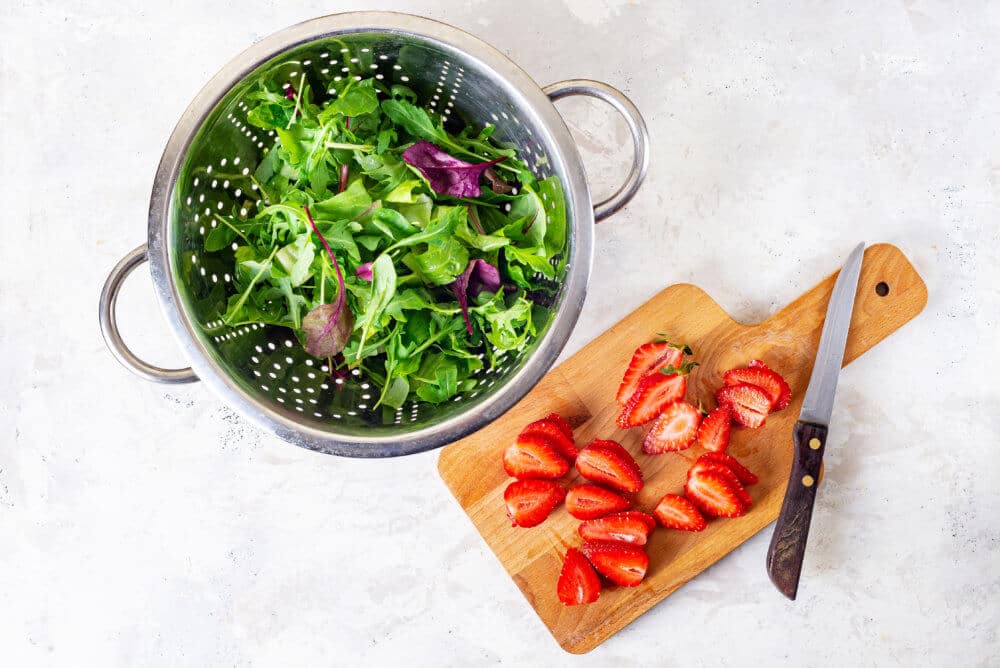 Greens in a sieve and chopped strawberries on a wooden cutting board with a knife.