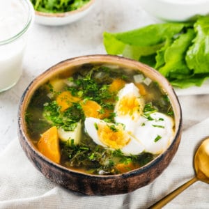 sorrel-soup-in-a-brown-bowl-on-a-white-towel-with-a-gold-spoon-some-sorrel-leaves-and-sour-cream-in-a-glass-in-the-background