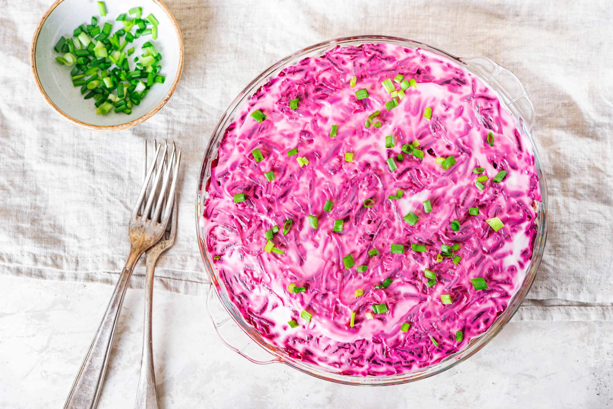 herring-salad-in-a-glass-dish-on-a-white-towel-with-forks-and-a-white-bowl-with-green-onions-on-the-side