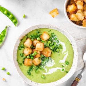 pea-soup-in-a-white-bowl-with-croutons-peas-and-dill-on-top-with-croutons-in-a-bowl-with-a-white-towel-a-spoon-and-peas-on-the-side