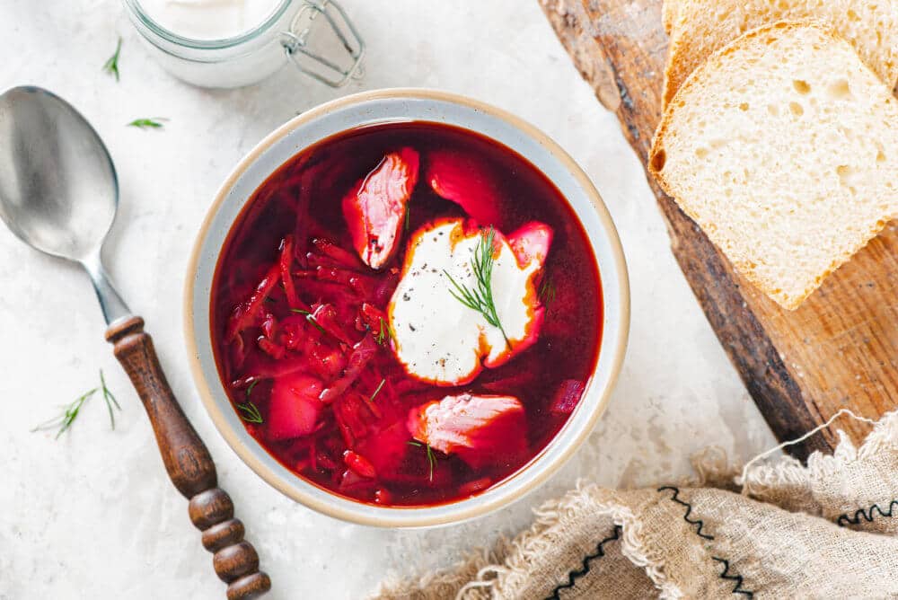 wooden-board-white-bread-white-bowl-red-borscht-sour-cream-dill-clear-bowl-with-sour-cream-and-a-wooden-spoon-brown-towel