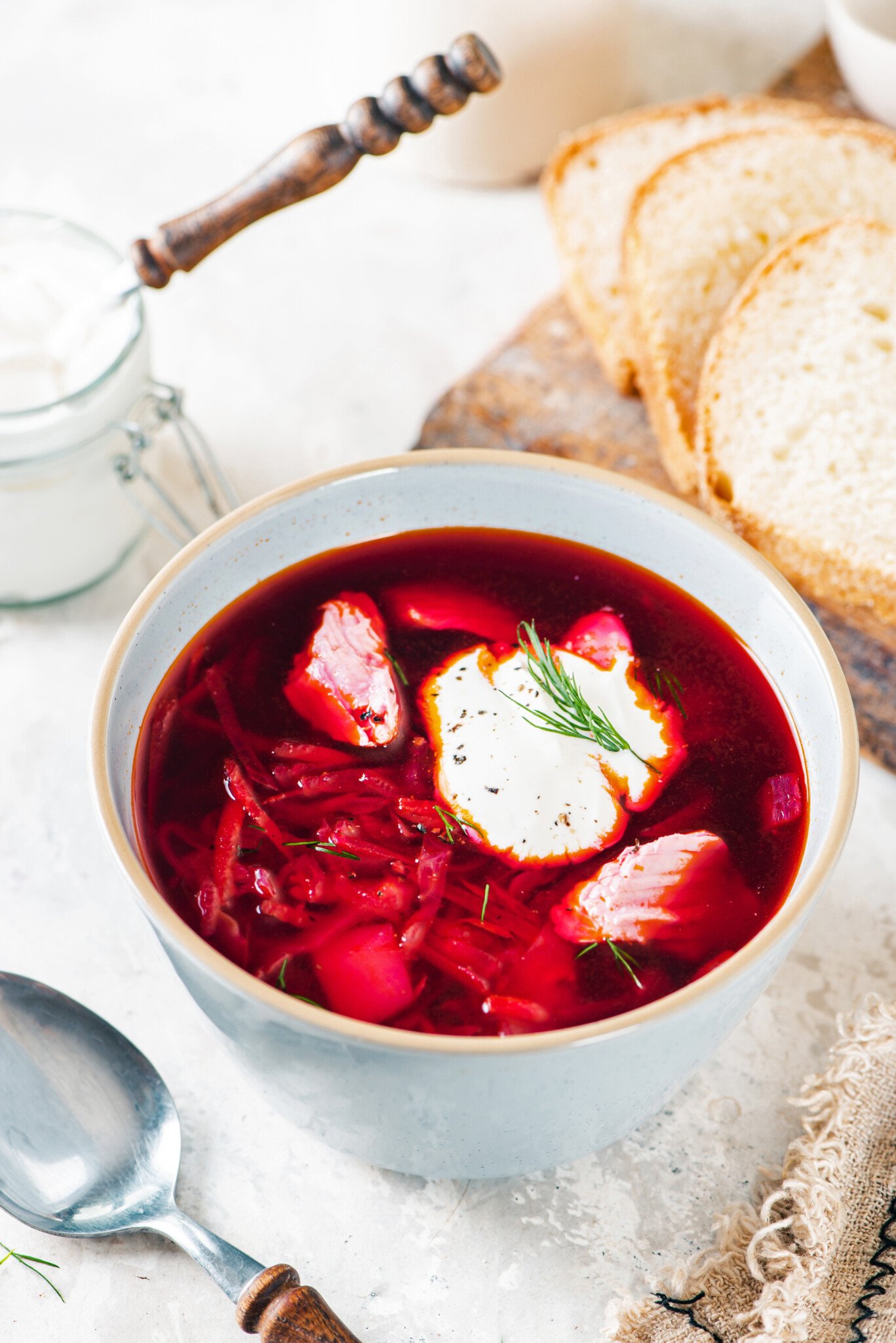 wooden-board-white-bread-white-bowl-red-borscht-sour-cream-dill-clear-bowl-with-sour-cream-and-a-wooden-spoon-brown-towel