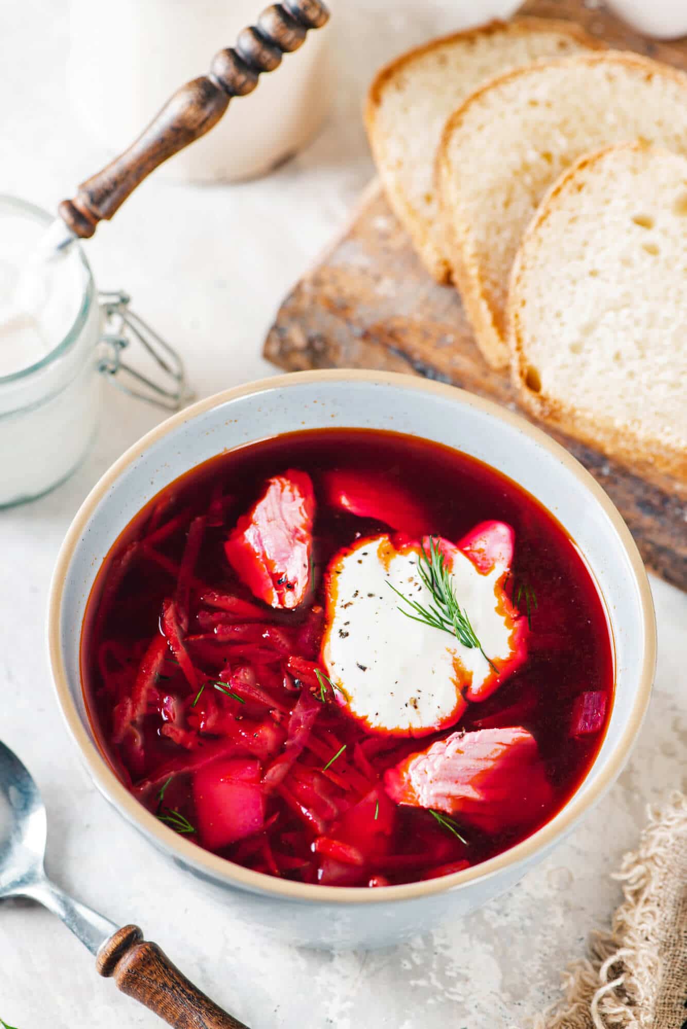 wooden-board-white-bread-white-bowl-red-borscht-sour-cream-dill-clear-bowl-with-sour-cream-and-a-wooden-spoon-brown-towel