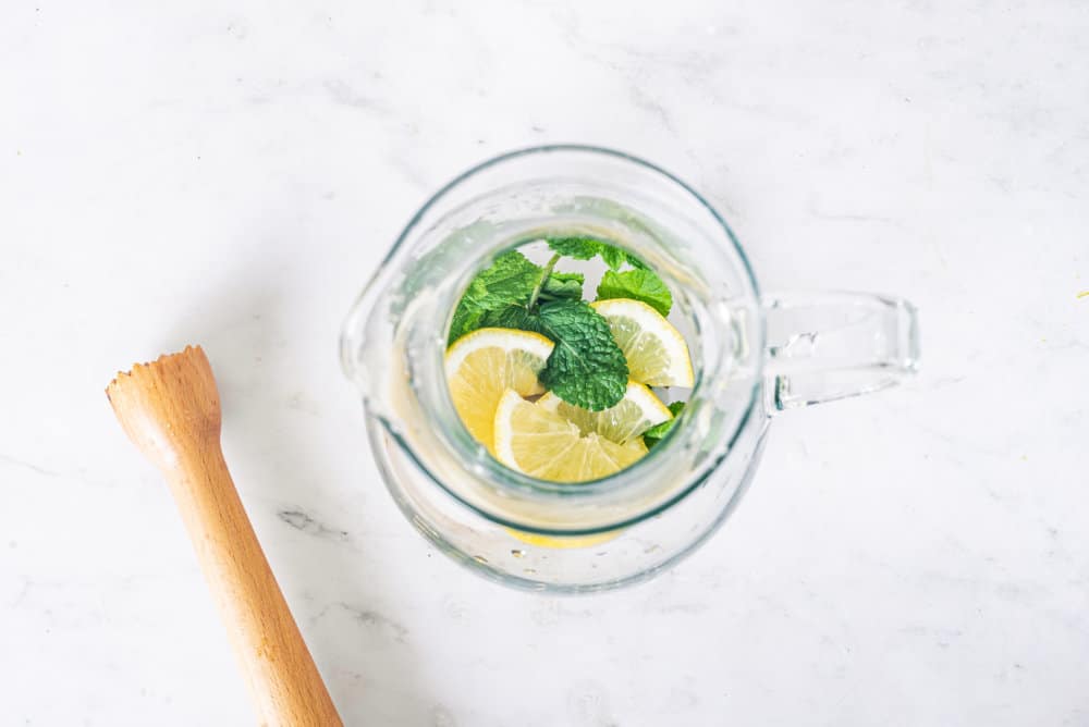 Lemon mint and ice in a glass pitcher with a wooden masher on the side.