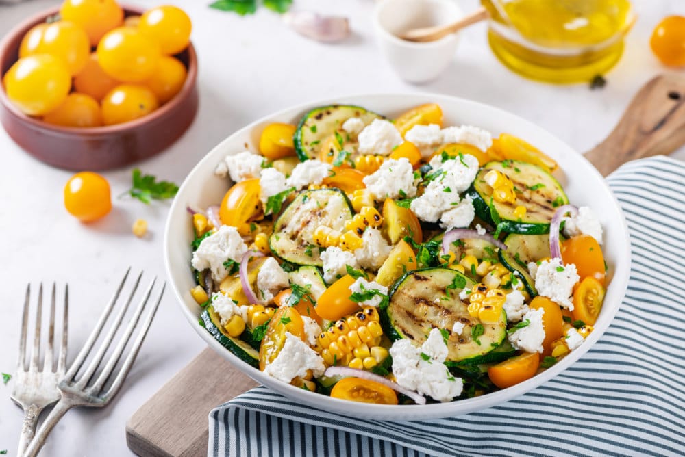 zucchini-corn-salad-in-a-white-bowl-on-a-wooden-board-with-a-blue-towel-with-forks-on-the-side-and-ingredients-in-the-background