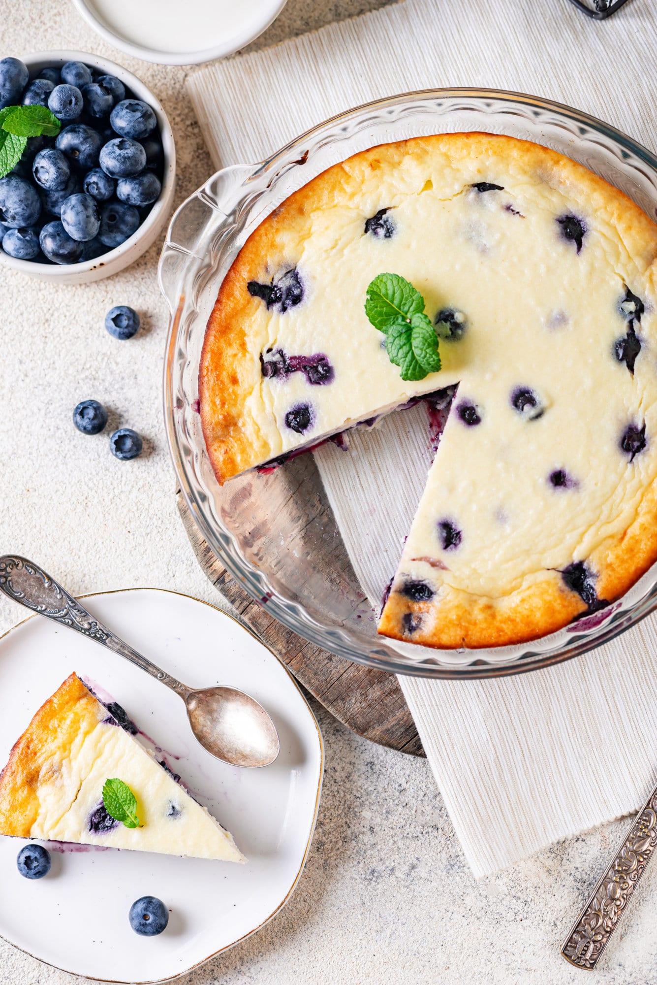 Blueberry cheese cake in a glass tray with a slice of cake on the side on a plate with a spoon.