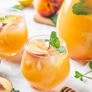 Peach lemonade in a glass cup with a pitcher on the side and fresh peaches around.