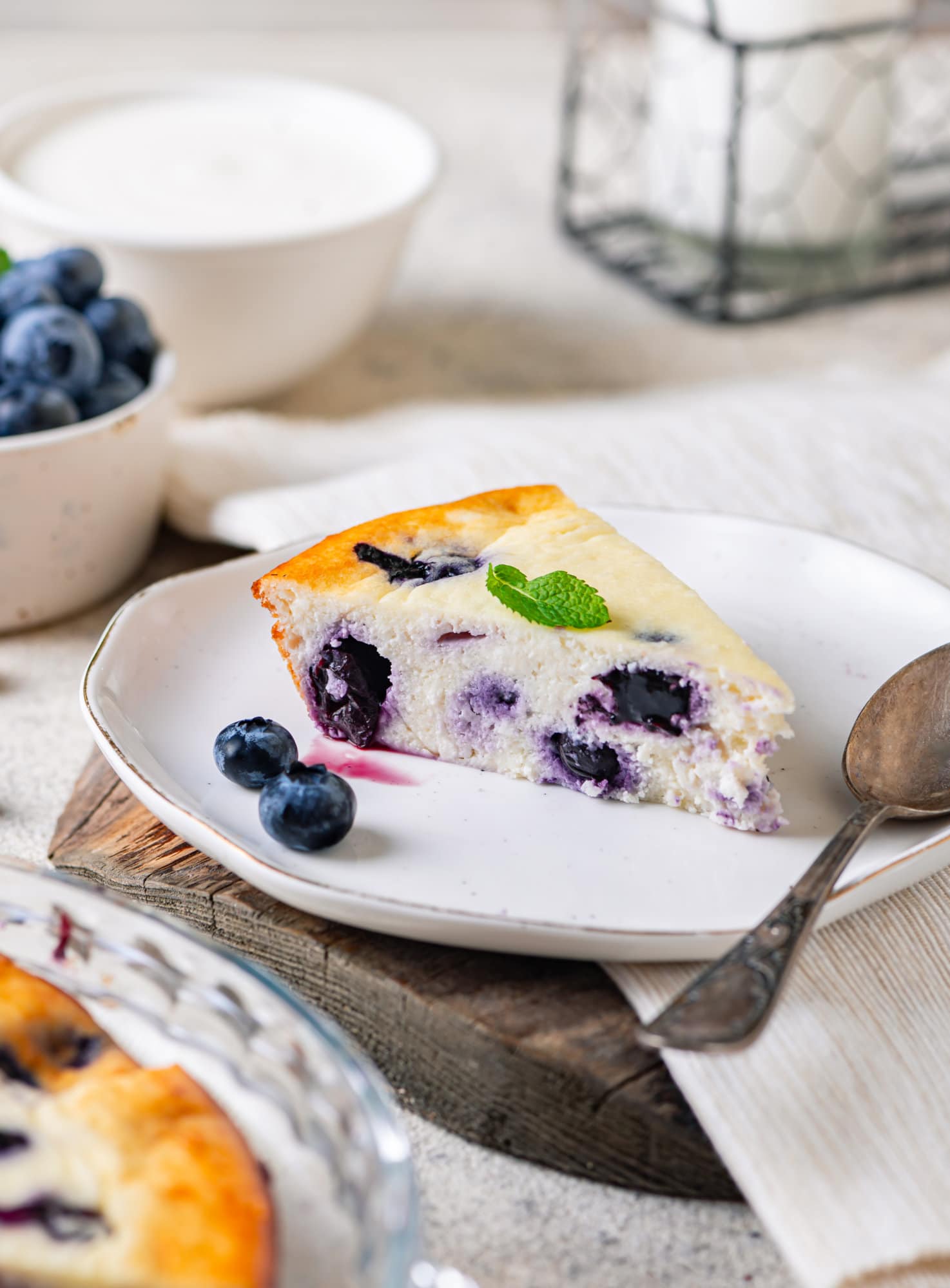 A slice of blueberry cheese cake on a white plate with a spoon and fresh blueberries on the plate. 