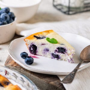 blueberry-cake-slice-on-a-white-plate-with-a-spoon-on-a-wooden-board-with-blueberries-on-the-plate