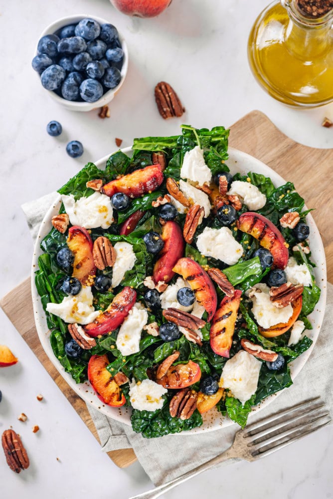 grilled-peaches-kale-salad-on-a-white-plate-on-a-wooden-board-on-a-grey-towel-with-a-fork-on-the-side-and-ingredients-in-the-background