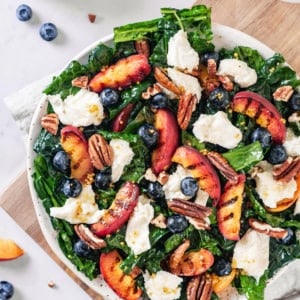 grilled-peaches-kale-salad-on-a-white-plate-on-a-wooden-board-on-a-grey-towel-with-a-fork-on-the-side-and-ingredients-in-the-background