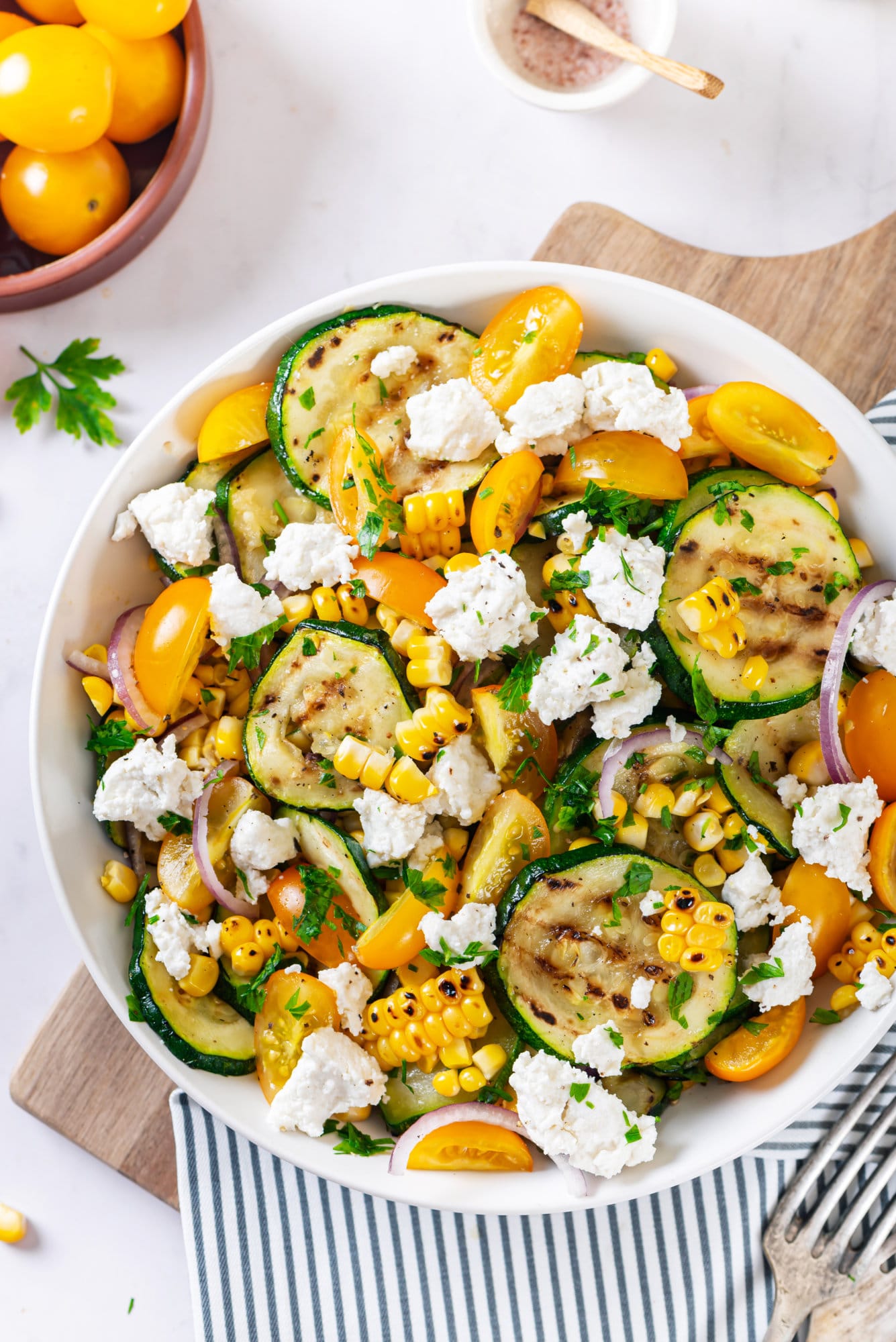 Zucchini corn salad in a white bowl on a blue striped towel on top of a wooden board surrounded by fresh ingredients for the salad. 