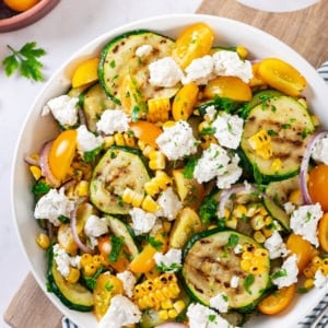 Zucchini corn salad in a white bowl on a blue striped towel on top of a wooden board surrounded by fresh ingredients for the salad.