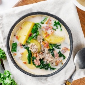 zuppa-toscana-in-a-grey-bowl-on-a-white-towel-on-a-wooden-board-with-a-spoon-and-kale-on-the-side