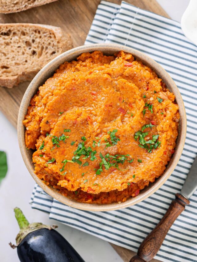 eggplant spread in a brown bowl on a blue towel with a wooden board, a towel, and a slice of bread on the side.
