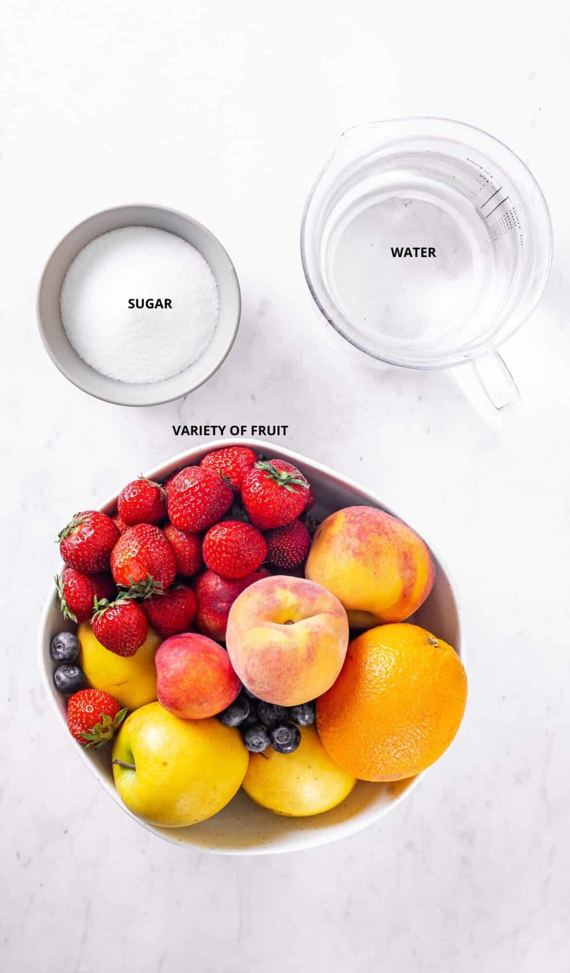 fruit juice ingredients, bowl of white sugar, pitcher of water and a bowl of variety of fresh fruit on a white marble background.